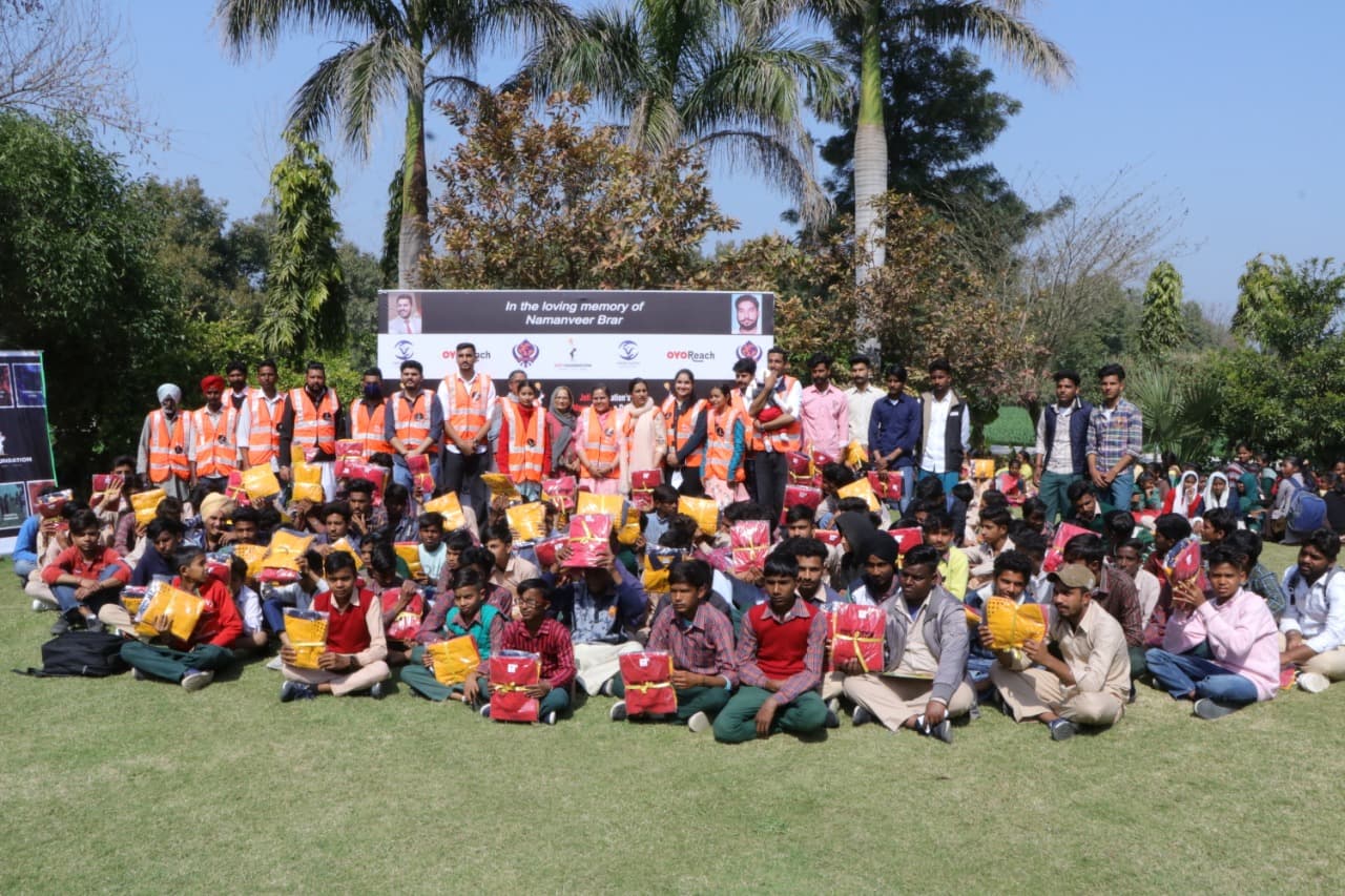 Joti Foundation volunteers in hi-vis vests standing behind rows of schoolchildren seated on grass holding relief kits at a distribution camp