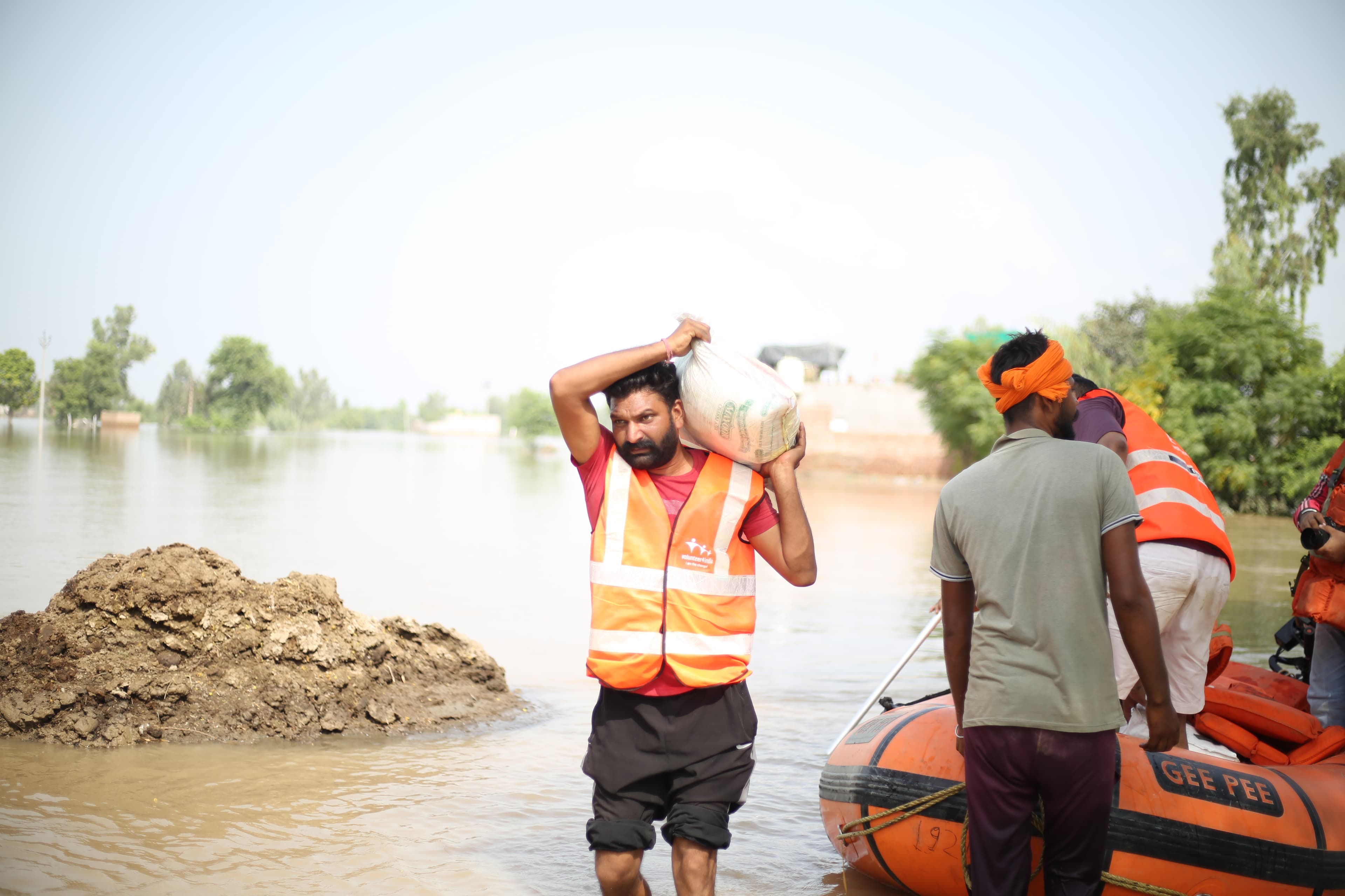 A relief worker in an orange vest wades through floodwater carrying a supply sack, with a rescue boat crew close behind