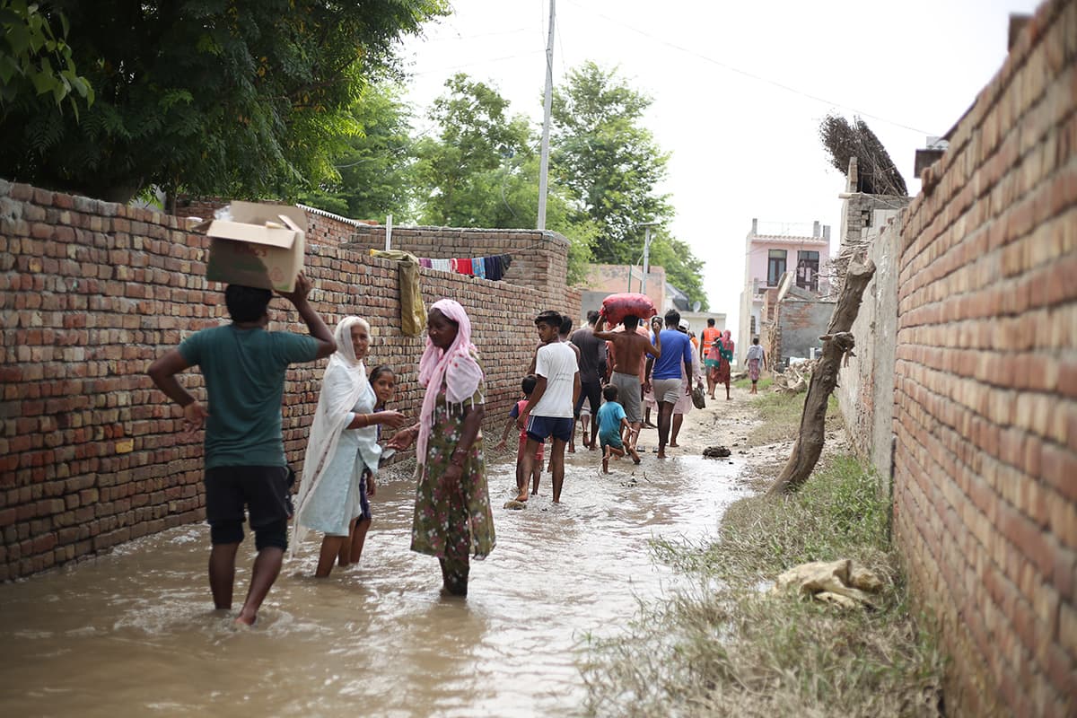 Villagers carrying boxes and bundles on their heads as they walk out through a flooded brick-walled lane